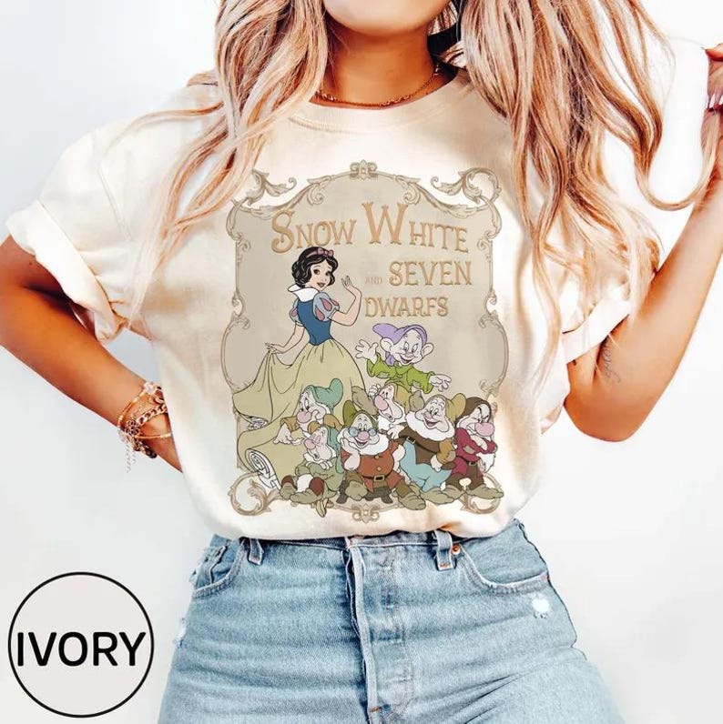 May include: Ivory-colored t-shirt featuring a graphic of Snow White and the Seven Dwarfs. The design includes an illustration of Snow White in a blue and yellow dress, surrounded by the seven dwarfs. The text "Snow White and Seven Dwarfs" is displayed above the characters.