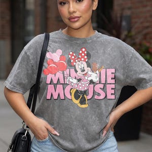 May include: Gray t-shirt featuring a Minnie Mouse graphic. The graphic includes Minnie Mouse holding balloons and a musical instrument, with the words "MINNIE MOUSE" in pink. The shirt has a vintage, washed look.