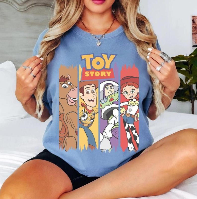 May include: A light blue t-shirt featuring the words "TOY STORY" in yellow and orange. The shirt displays cartoon characters from the movie, including Woody, Buzz Lightyear, and Jessie, set against colorful vertical stripes.