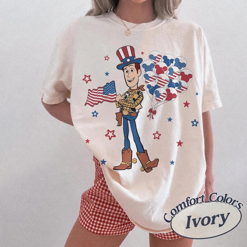May include: Ivory-coloured t-shirt featuring a cartoon cowboy holding an American flag and balloons. The design includes red, white, and blue stars and balloons. The shirt has the words "Comfort Colors Ivory" printed on it.