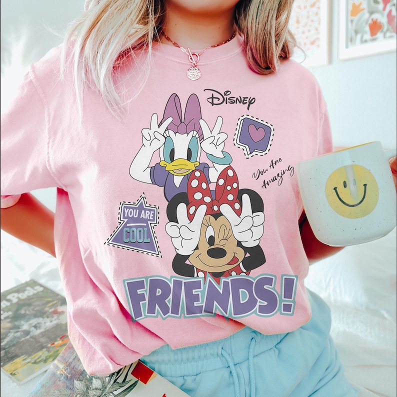 Może przedstawiać: R&oacute;żowa koszulka z grafiką Kaczki Daisy i Myszki Minnie z Disneya pokazujących znak pokoju. Słowo "FRIENDS!" jest w fioletowych literach blokowych. Widoczne jest logo Disneya, a także frazy "You Are Cool" i "You Are Amazing".