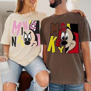 Op de afbeelding: Twee T-shirts, &eacute;&eacute;n beige en &eacute;&eacute;n bruin, met cartoonkarakterontwerpen. Het beige shirt toont "MINNIE" in roze en geel met een Minnie Mouse-afbeelding. Het bruine shirt heeft "MICKEY" in bruin en geel met een Mickey Mouse-afbeelding.