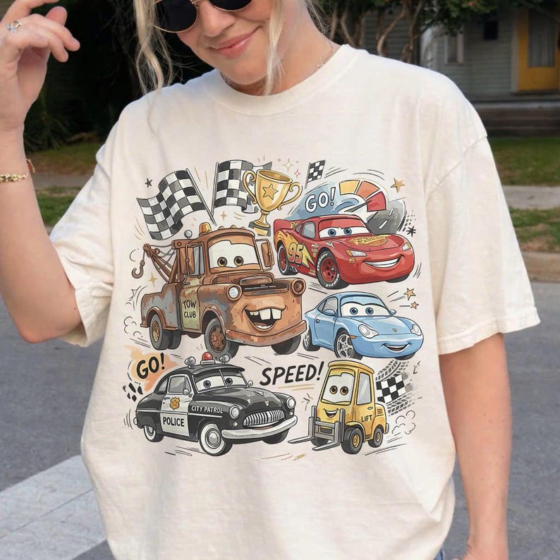 May include: A cream-colored t-shirt featuring a graphic print of characters from the movie Cars, including a brown tow truck, a red race car, a blue sports car, a black police car, and a yellow forklift, with checkered flags and the text Go and Speed.