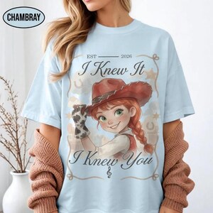 May include: A light blue t-shirt featuring a graphic of a cartoon character wearing a brown cowboy hat and a cow-print glove. The text reads "EST 2026 I Knew It I Knew You" with a musical note symbol. The design is framed by a rope-like border.