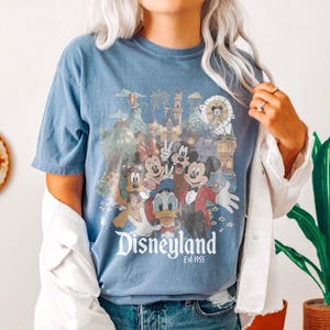 Puede incluir: Camiseta azul con un dise&ntilde;o de Disneyland que presenta a Mickey Mouse, Minnie Mouse, Donald Duck y Goofy. El dise&ntilde;o incluye un castillo, fuegos artificiales y el texto "Disneyland Est. 1955". La camiseta tiene mangas cortas.