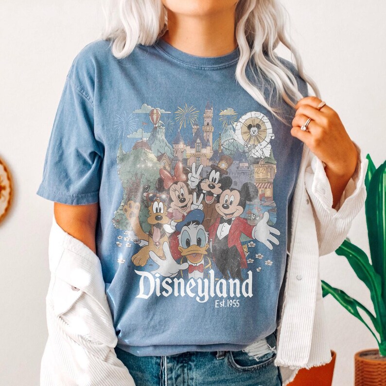 Puede incluir: Camiseta azul con un dise&ntilde;o de Disneyland que presenta a Mickey Mouse, Minnie Mouse, Donald Duck y Goofy. El dise&ntilde;o incluye un castillo, fuegos artificiales y el texto "Disneyland Est. 1955". La camiseta tiene mangas cortas.
