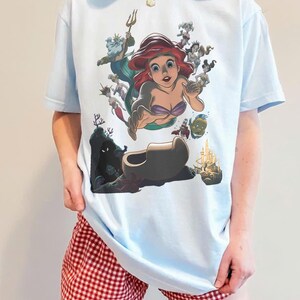 May include: A light blue t-shirt featuring a graphic print of Ariel from The Little Mermaid, surrounded by various underwater characters including King Triton, Flounder, and Sebastian, with a castle and dark cave depicted at the bottom.