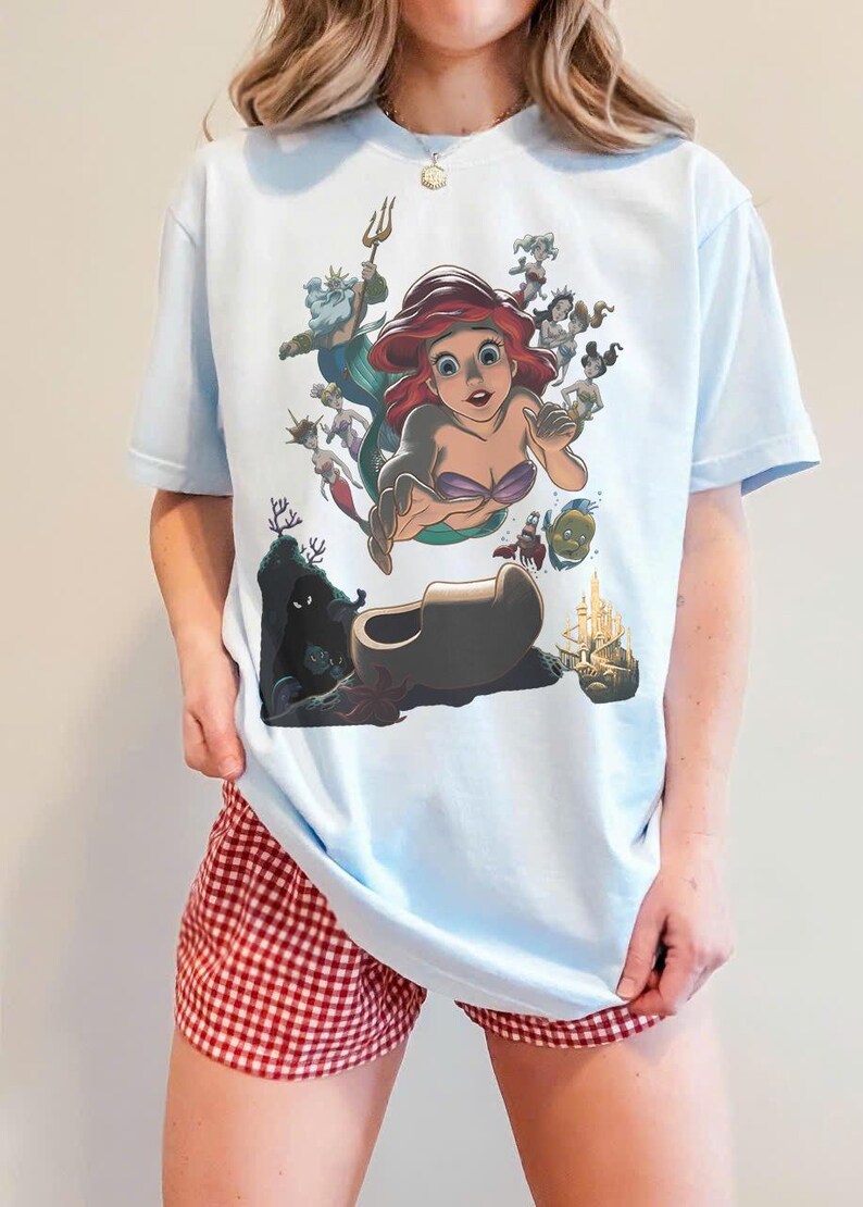 May include: A light blue t-shirt featuring a graphic print of Ariel from The Little Mermaid, surrounded by various underwater characters including King Triton, Flounder, and Sebastian, with a castle and dark cave depicted at the bottom.