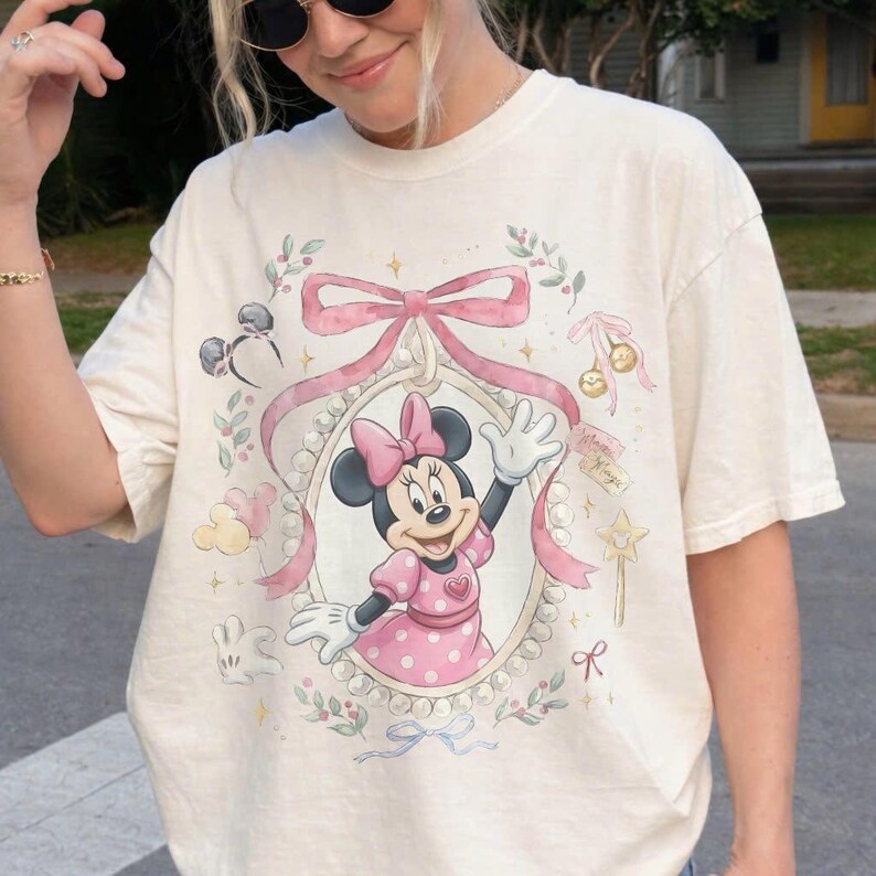 May include: A cream-colored short-sleeve t-shirt featuring a graphic of Minnie Mouse inside a pearl-bordered oval frame. The design includes a large pink bow, small floral sprigs, a star wand, and a pair of mouse ears, all in soft pastel tones.