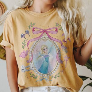 May include: A mustard yellow short-sleeve t-shirt featuring a graphic print of a blonde character in a blue dress inside a pearl-bordered oval frame. The design includes a large pink bow, Mickey Mouse ears, white gloves, and a star-shaped wand.
