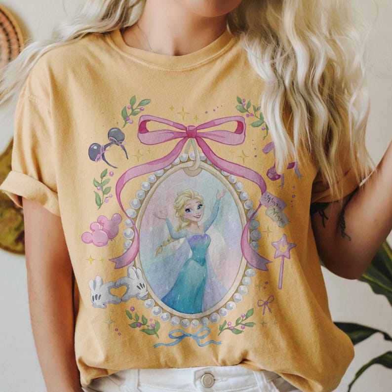 May include: A mustard yellow short-sleeve t-shirt featuring a graphic print of a blonde character in a blue dress inside a pearl-bordered oval frame. The design includes a large pink bow, Mickey Mouse ears, white gloves, and a star-shaped wand.