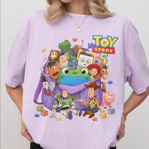 May include: A light purple t-shirt featuring a graphic print of Toy Story 5 characters including Woody, Buzz Lightyear, Rex, Slinky Dog, Mr. Potato Head, Jessie, Bullseye, Hamm, and Forky emerging from a purple backpack with heart patterns.