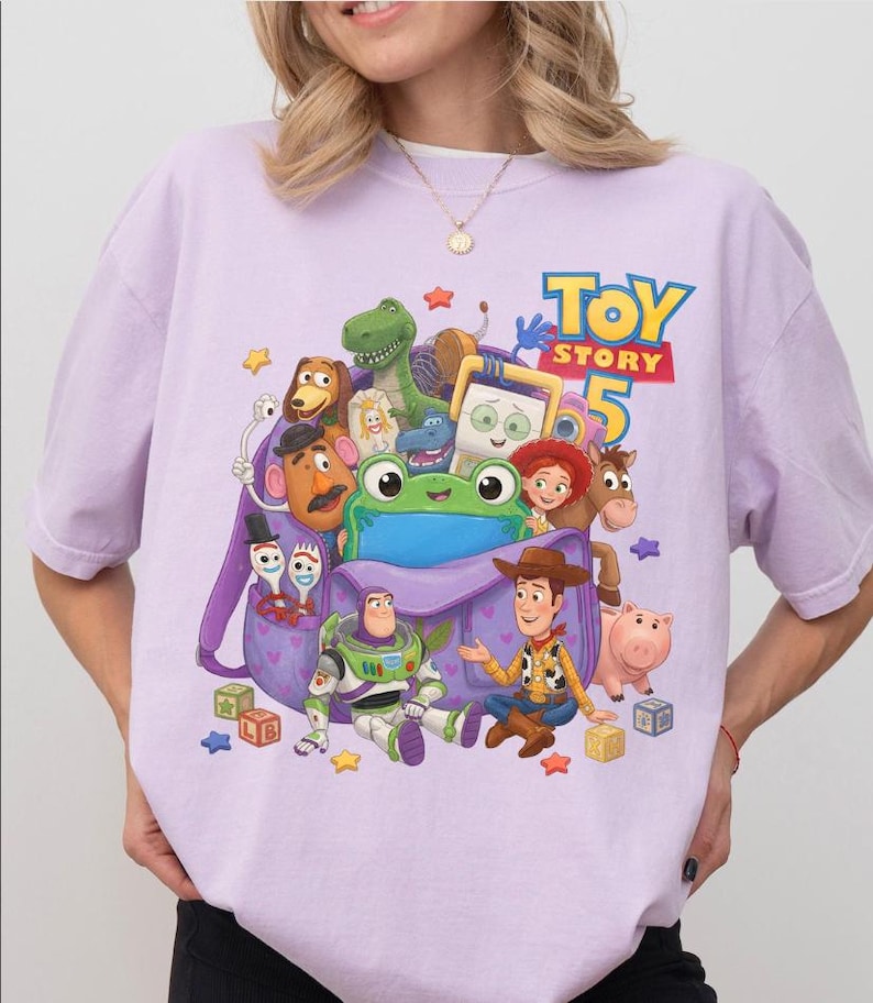 May include: A light purple t-shirt featuring a graphic print of Toy Story 5 characters including Woody, Buzz Lightyear, Rex, Slinky Dog, Mr. Potato Head, Jessie, Bullseye, Hamm, and Forky emerging from a purple backpack with heart patterns.
