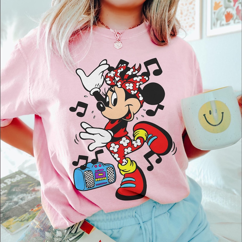 Pu&ograve; includere: Maglietta rosa chiaro con una grafica di Minnie Mouse che balla con note musicali e un boombox. Minnie Mouse indossa un completo floreale rosso e bianco e scarpe rosse. Viene tenuta una tazza bianca con una faccina gialla.