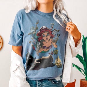 May include: A blue short-sleeve t-shirt featuring a graphic print of the character Ariel from The Little Mermaid, surrounded by various supporting characters, a castle, and underwater elements. The design is centered on the front of the shirt.