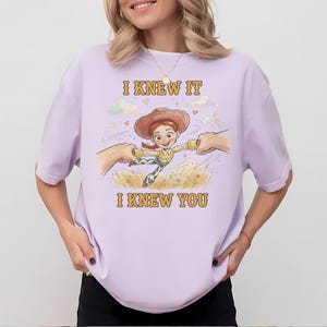 May include: A light purple t-shirt featuring a graphic illustration of a cartoon cowgirl character with red hair and a brown hat, being held by two hands. The text "I KNEW IT" and "I KNEW YOU" is printed in yellow above and below the character.