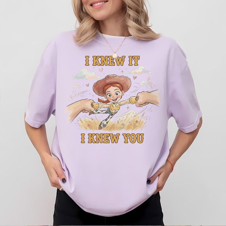 May include: A light purple t-shirt featuring a graphic illustration of a cartoon cowgirl character with red hair and a brown hat, being held by two hands. The text "I KNEW IT" and "I KNEW YOU" is printed in yellow above and below the character.