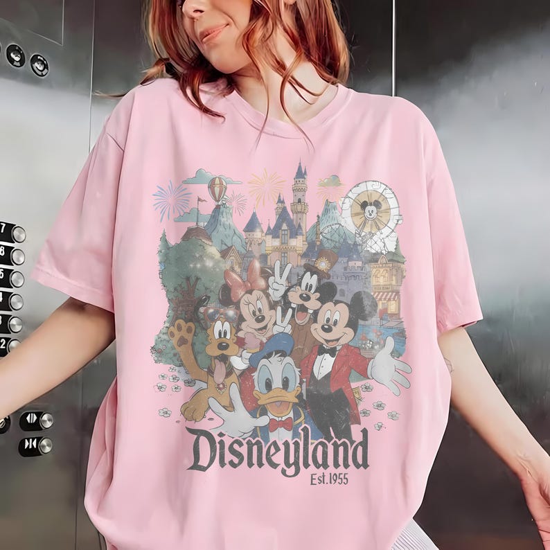 Puede incluir: Camiseta rosa claro con un dise&ntilde;o de Disneyland que presenta a Mickey, Minnie, Donald y Pluto. El dise&ntilde;o incluye un castillo, fuegos artificiales y una noria. El texto "Disneyland Est. 1955" est&aacute; impreso debajo de los personajes.