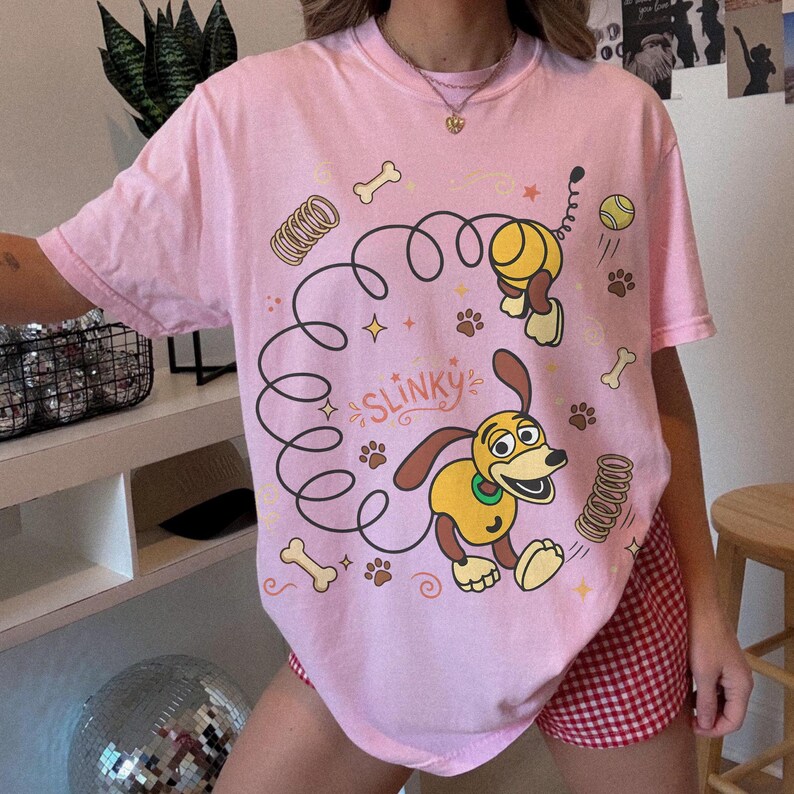 May include: A light pink t-shirt featuring a cartoon image of Slinky Dog from Toy Story. The design includes Slinky Dog, a ball, paw prints, and the word "Slinky". The shirt is a crew neck and has short sleeves.