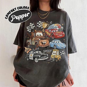 May include: A charcoal gray t-shirt featuring a graphic print of various animated car characters, including a brown tow truck, a red race car, a blue sports car, a black police car, and a yellow forklift, with the text COMFORT COLORS Pepper.