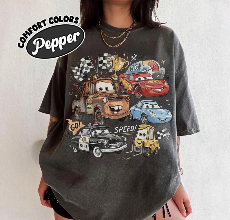May include: A charcoal gray t-shirt featuring a graphic print of various animated car characters, including a brown tow truck, a red race car, a blue sports car, a black police car, and a yellow forklift, with the text COMFORT COLORS Pepper.