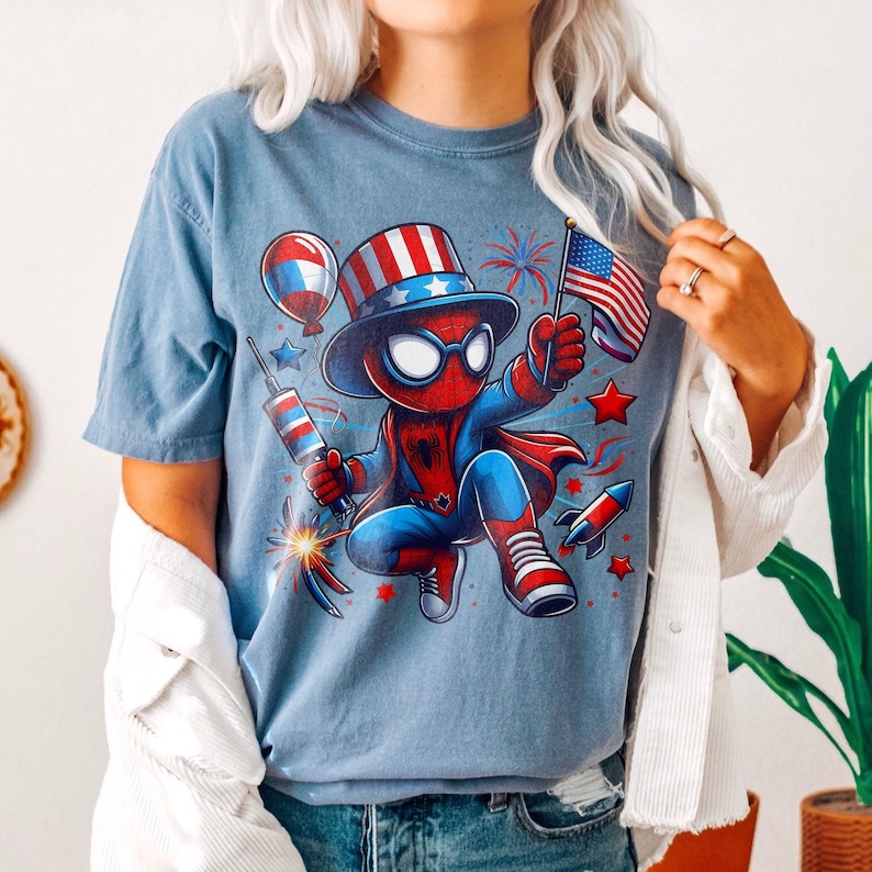 K&ouml;nnte beinhalten: Hellblaues T-Shirt mit einer Cartoon-Figur von Spider-Man in einem patriotischen Outfit. Die Figur h&auml;lt eine kleine amerikanische Flagge und einen Feuerwerksk&ouml;rper, mit Feuerwerk und Sternen drumherum. Das Design umfasst rote, wei&szlig;e und blaue Farben.