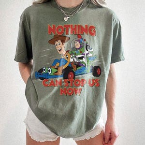 以下が含まれることがあります： オリーブグリーンのTシャツ。ウッディとバズ・ライトイヤーがレーシングカーに乗ったグラフィックと、赤字で「NOTHING CAN STOP US NOW」の文字がプリントされています。ゆったりとしたフィット感で、半袖です。