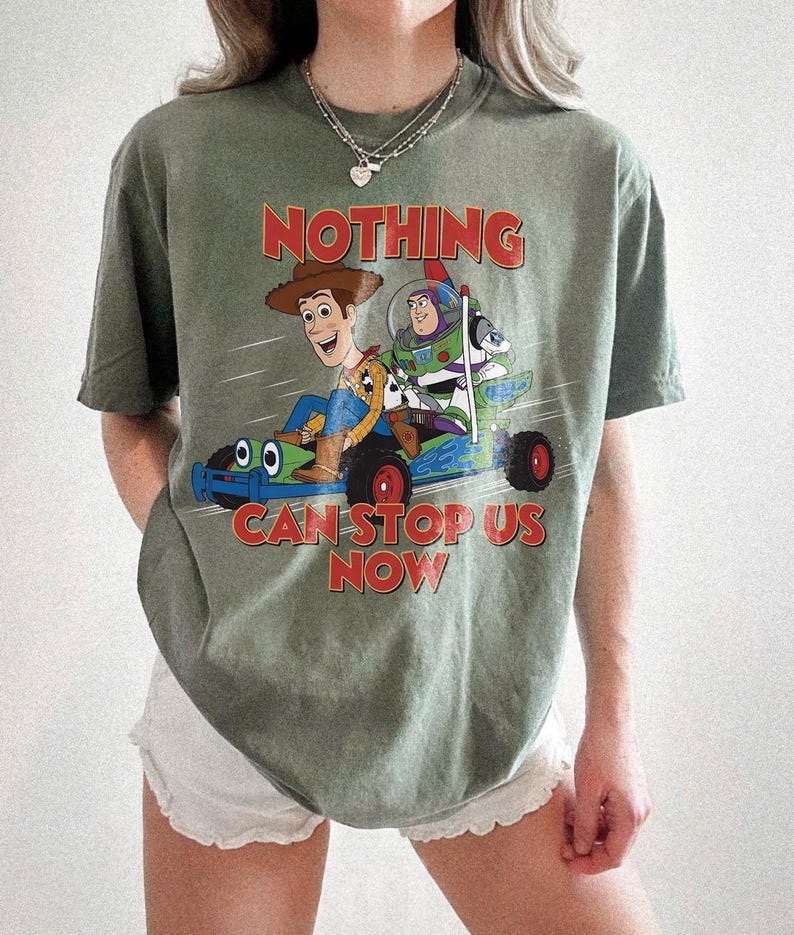 以下が含まれることがあります： オリーブグリーンのTシャツ。ウッディとバズ・ライトイヤーがレーシングカーに乗ったグラフィックと、赤字で「NOTHING CAN STOP US NOW」の文字がプリントされています。ゆったりとしたフィット感で、半袖です。