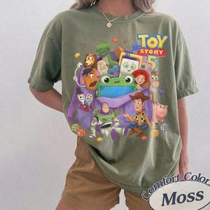 May include: An olive green crew neck t-shirt featuring a colorful graphic print of various Toy Story characters, including Woody, Buzz Lightyear, and a large green frog, with the text Toy Story 5 printed in yellow and blue above the characters.