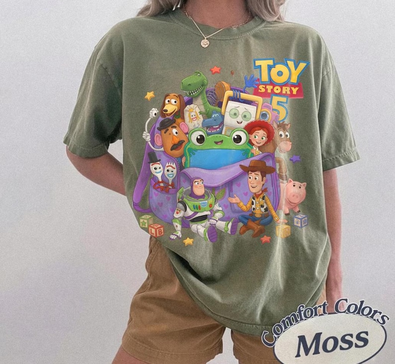 May include: An olive green crew neck t-shirt featuring a colorful graphic print of various Toy Story characters, including Woody, Buzz Lightyear, and a large green frog, with the text Toy Story 5 printed in yellow and blue above the characters.