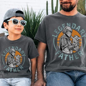 K&ouml;nnte beinhalten: Zwei anthrazitfarbene T-Shirts mit dem Text "LEGENDARY FATHER" und einer Grafik eines Mandalorianer-Charakters, der Grogu h&auml;lt. Die Shirts werden von einem Kind und einem Erwachsenen getragen, beide mit Sonnenbrille und Kappe.