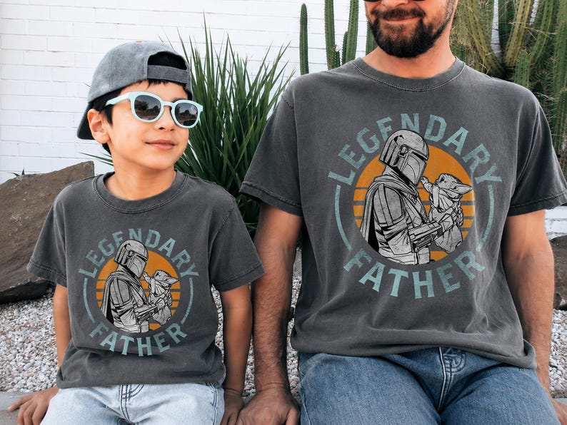 K&ouml;nnte beinhalten: Zwei anthrazitfarbene T-Shirts mit dem Text "LEGENDARY FATHER" und einer Grafik eines Mandalorianer-Charakters, der Grogu h&auml;lt. Die Shirts werden von einem Kind und einem Erwachsenen getragen, beide mit Sonnenbrille und Kappe.