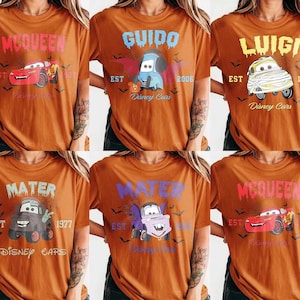 May include: Six orange t-shirts featuring Disney Cars characters. Each shirt has a different character name in a spooky font, with the character's image, the word "EST", and a year. The shirts are a Halloween theme.