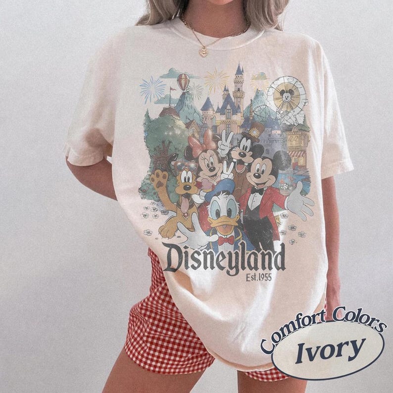 Puede incluir: Camiseta color marfil con un dise&ntilde;o de Disneyland que presenta a Mickey Mouse, Minnie Mouse, Donald Duck y Goofy. El dise&ntilde;o incluye un castillo, fuegos artificiales y una noria. El texto "Disneyland Est. 1955" est&aacute; impreso debajo.