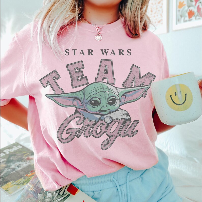 Puede incluir: Una camiseta de manga corta de color rosa claro con un gr&aacute;fico del personaje Grogu de Star Wars. El texto dice STAR WARS TEAM GROGU en una fuente desgastada. La camiseta se combina con pantalones azul claro y una taza blanca con una cara sonriente.