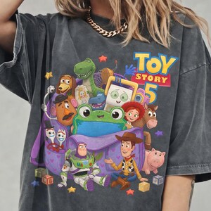 May include: A charcoal gray short-sleeve t-shirt featuring a graphic print of Toy Story 5 characters including Woody, Buzz Lightyear, Rex, Slinky Dog, Mr. Potato Head, Jessie, Bullseye, Hamm, and Forky emerging from a purple backpack.