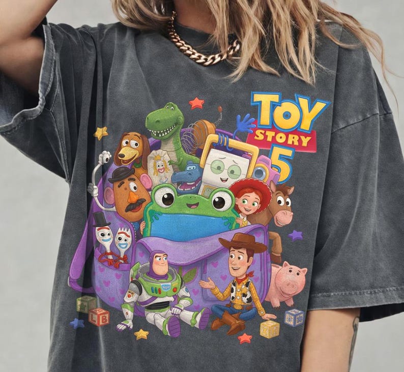 May include: A charcoal gray short-sleeve t-shirt featuring a graphic print of Toy Story 5 characters including Woody, Buzz Lightyear, Rex, Slinky Dog, Mr. Potato Head, Jessie, Bullseye, Hamm, and Forky emerging from a purple backpack.