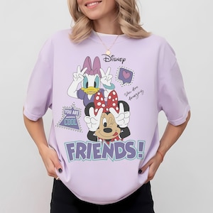 Może przedstawiać: Jasnofioletowa koszulka z Myszką Minnie i Daisy Duck pokazującymi znak pokoju. Koszulka Disneya ma fioletowy napis "FRIENDS!" oraz frazy "You Are Cool" i "You Are Amazing".