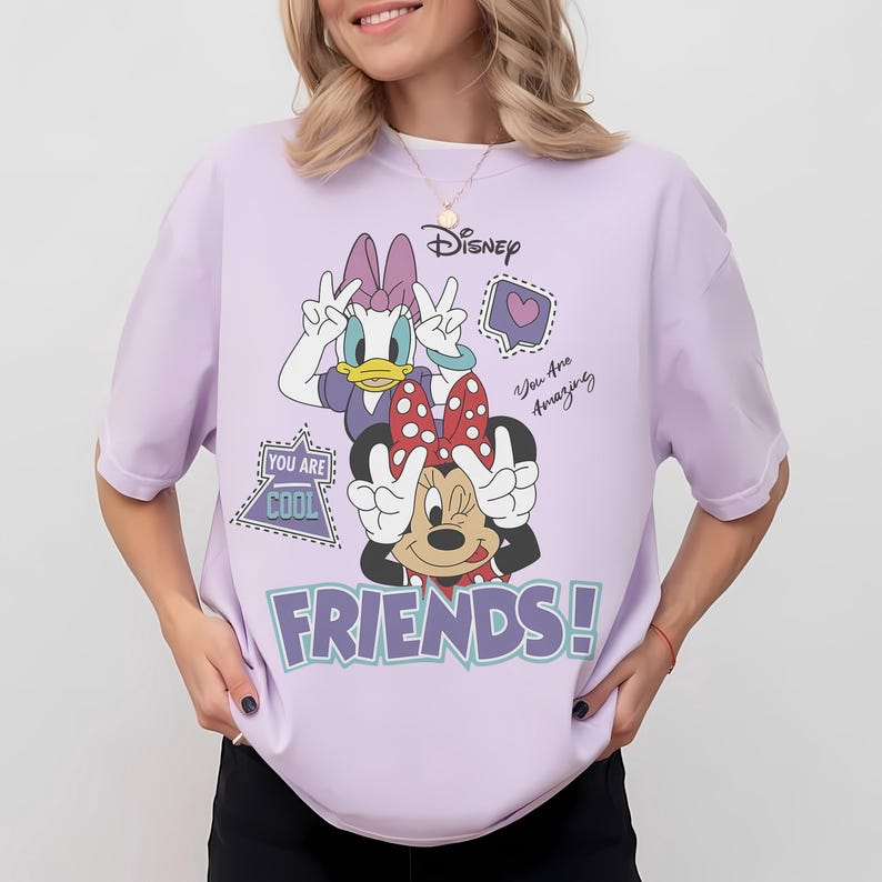 Może przedstawiać: Jasnofioletowa koszulka z Myszką Minnie i Daisy Duck pokazującymi znak pokoju. Koszulka Disneya ma fioletowy napis "FRIENDS!" oraz frazy "You Are Cool" i "You Are Amazing".