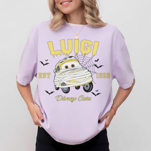 May include: A light purple t-shirt featuring a cartoon image of Luigi from Disney Cars wrapped in bandages. The text "LUIGI", "EST 1959", and "Disney Cars" are printed in yellow. Small black bats are scattered around the design.