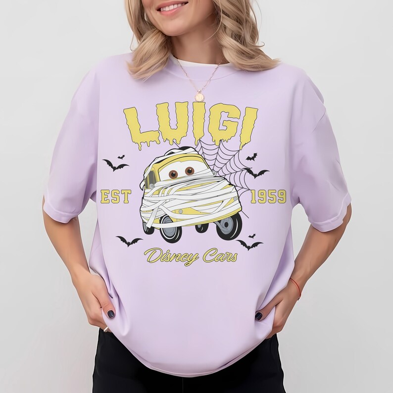 May include: A light purple t-shirt featuring a cartoon image of Luigi from Disney Cars wrapped in bandages. The text "LUIGI", "EST 1959", and "Disney Cars" are printed in yellow. Small black bats are scattered around the design.