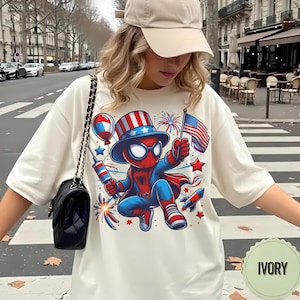 K&ouml;nnte beinhalten: Elfenbeinfarbenes T-Shirt mit einem Cartoon-Spider-Man-Charakter in patriotischer Kleidung, der eine US-Flagge und Feuerwerk h&auml;lt. Das Design umfasst rote, wei&szlig;e und blaue Elemente, Sterne und Raketen. Ein Freizeitkleidungsst&uuml;ck.