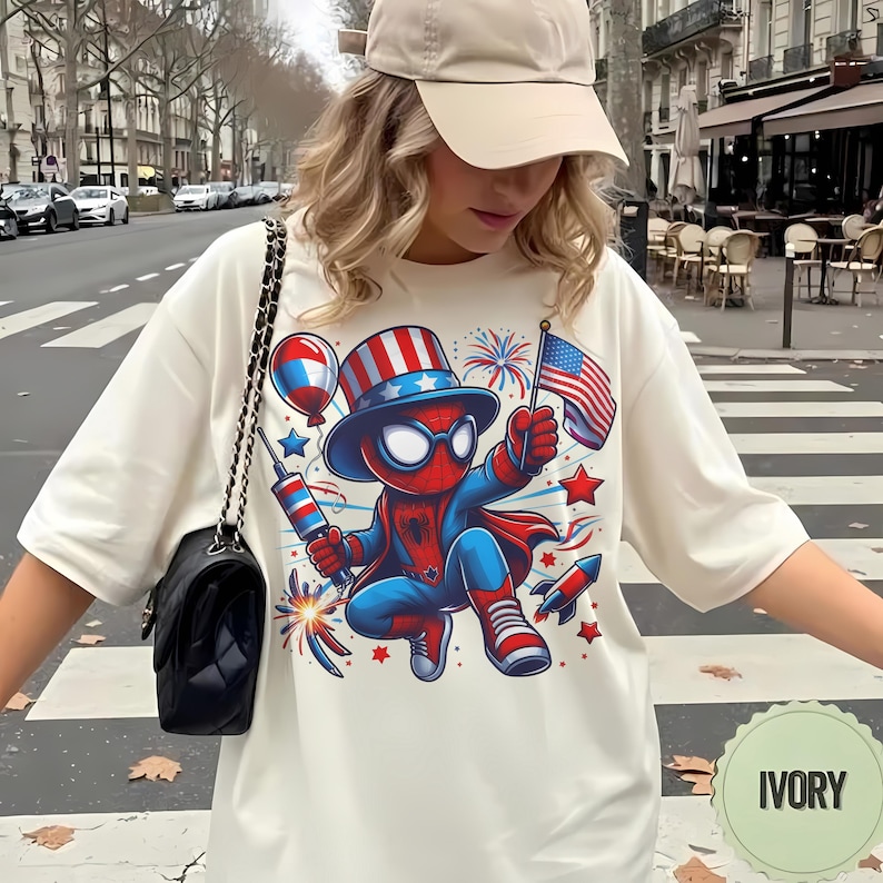 K&ouml;nnte beinhalten: Elfenbeinfarbenes T-Shirt mit einem Cartoon-Spider-Man-Charakter in patriotischer Kleidung, der eine US-Flagge und Feuerwerk h&auml;lt. Das Design umfasst rote, wei&szlig;e und blaue Elemente, Sterne und Raketen. Ein Freizeitkleidungsst&uuml;ck.