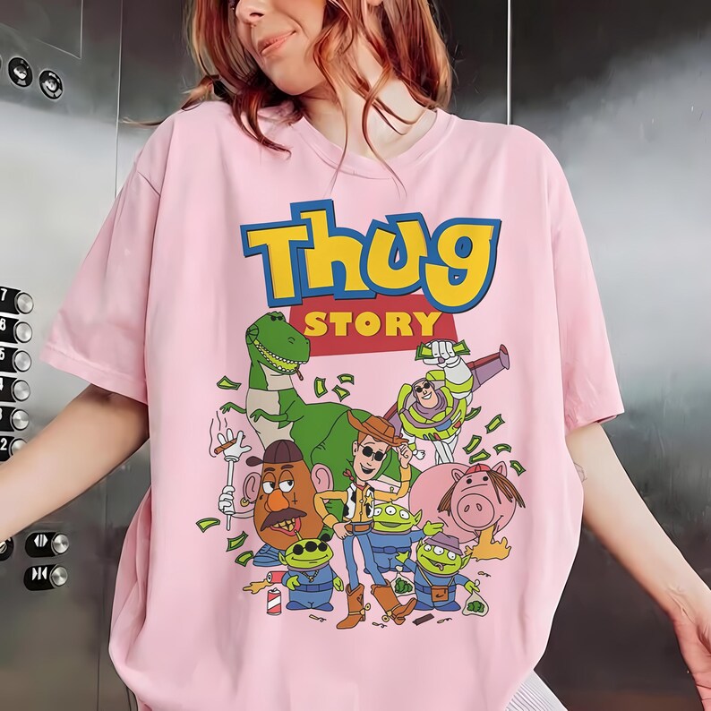 Pode incluir: T-shirt rosa claro com um gr&aacute;fico de desenho animado com o texto "Thug Story". O gr&aacute;fico inclui v&aacute;rios personagens animados, notas de dinheiro e outros elementos. A camisa tem mangas curtas e gola redonda.