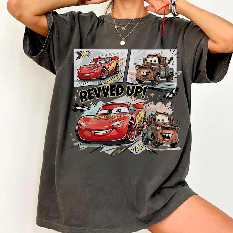 May include: A charcoal gray short-sleeve t-shirt featuring a graphic print of the characters Lightning McQueen and Mater from the movie Cars. The design includes the text REVVED UP! along with checkered flag patterns and stylized motion lines.