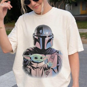 May include: Cream-colored t-shirt featuring a graphic of The Mandalorian and Grogu from the Star Wars series. The design includes the Mandalorian's helmet in shades of blue and Grogu in a tan robe, with a light blue skin tone.
