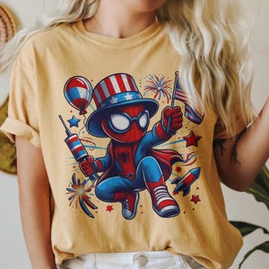 K&ouml;nnte beinhalten: Senfgelbes T-Shirt mit einer Cartoon-Figur von Spider-Man in einem patriotischen Outfit. Die Figur tr&auml;gt einen Zylinder und einen Umhang und h&auml;lt eine Flagge und ein Feuerwerk. Das Design verwendet rote, wei&szlig;e und blaue Akzente mit Feuerwerk und Sternen.