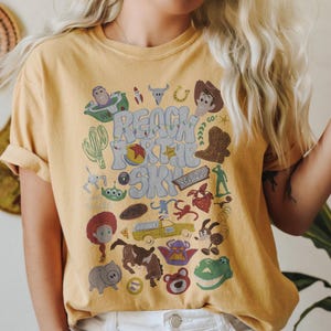 May include: A mustard yellow t-shirt featuring a graphic print with the text "REACH FOR THE SKY" surrounded by various cartoon character illustrations, including a cowboy, a space ranger, a green alien, a dinosaur, and a yellow truck.