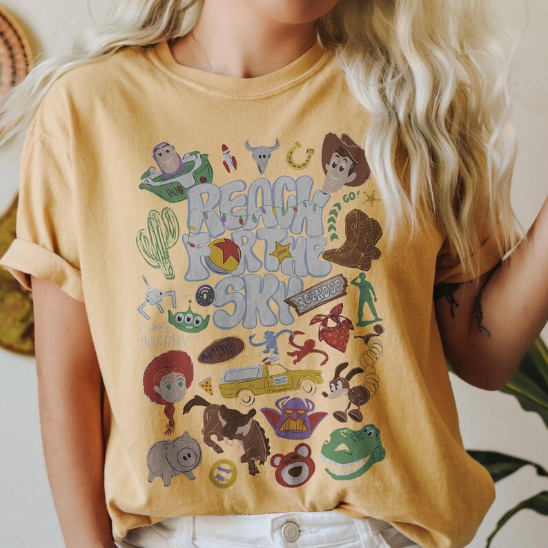 May include: A mustard yellow t-shirt featuring a graphic print with the text "REACH FOR THE SKY" surrounded by various cartoon character illustrations, including a cowboy, a space ranger, a green alien, a dinosaur, and a yellow truck.