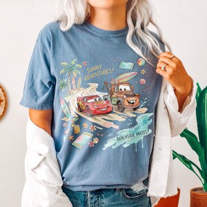 May include: A blue short-sleeve t-shirt featuring a graphic print of Lightning McQueen and Mater driving on a beach road. The design includes palm trees, surfboards, flowers, and the text Sunny Adventures and Beachside Mates in yellow and black.